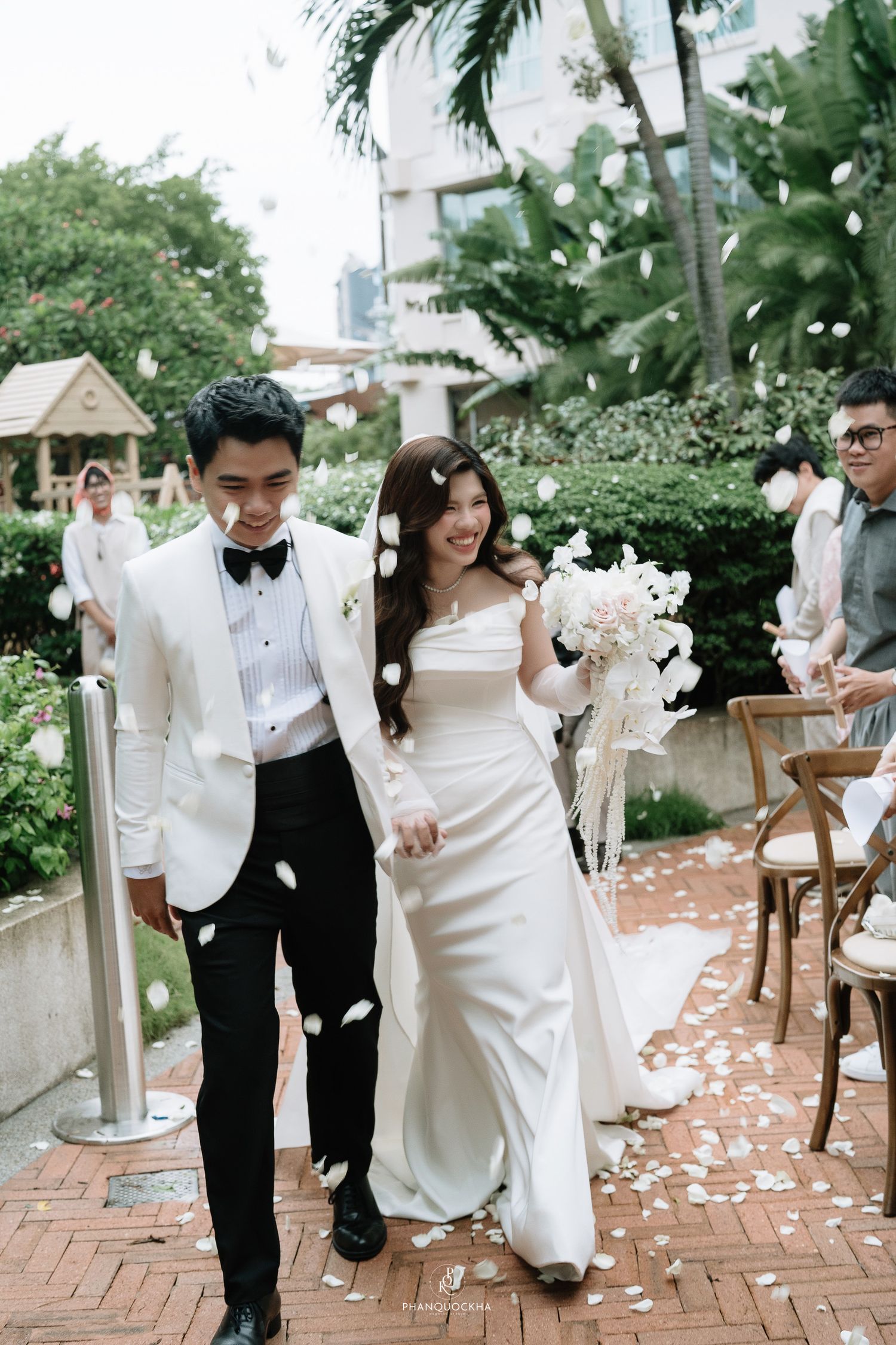 Happy newlyweds walk through petal toss on brick path after outdoor garden wedding ceremony, bride holding white bouquet.
