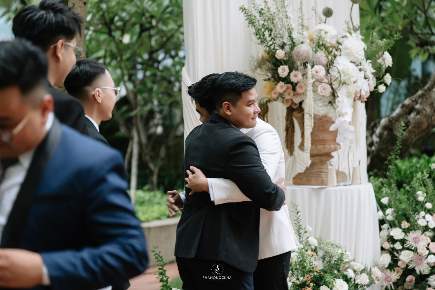 Two men share a warm embrace at an elegant outdoor wedding ceremony with floral decorations.