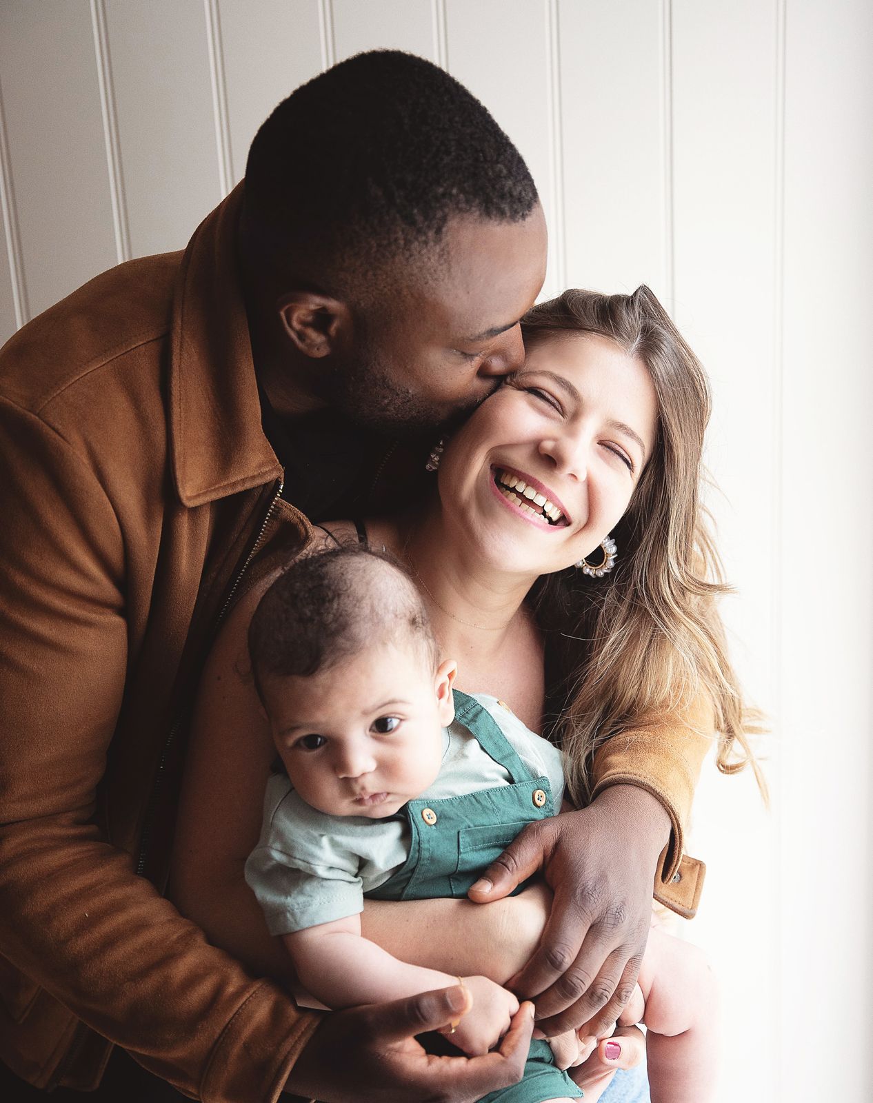 A couple in a tender family moment laugh and embrace with their baby wearing a teal outfit against a light backdrop. At a studio in Bexley