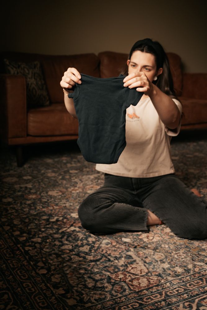 Someone sits cross-legged on a patterned rug holding up and examining a dark colored baby shirt