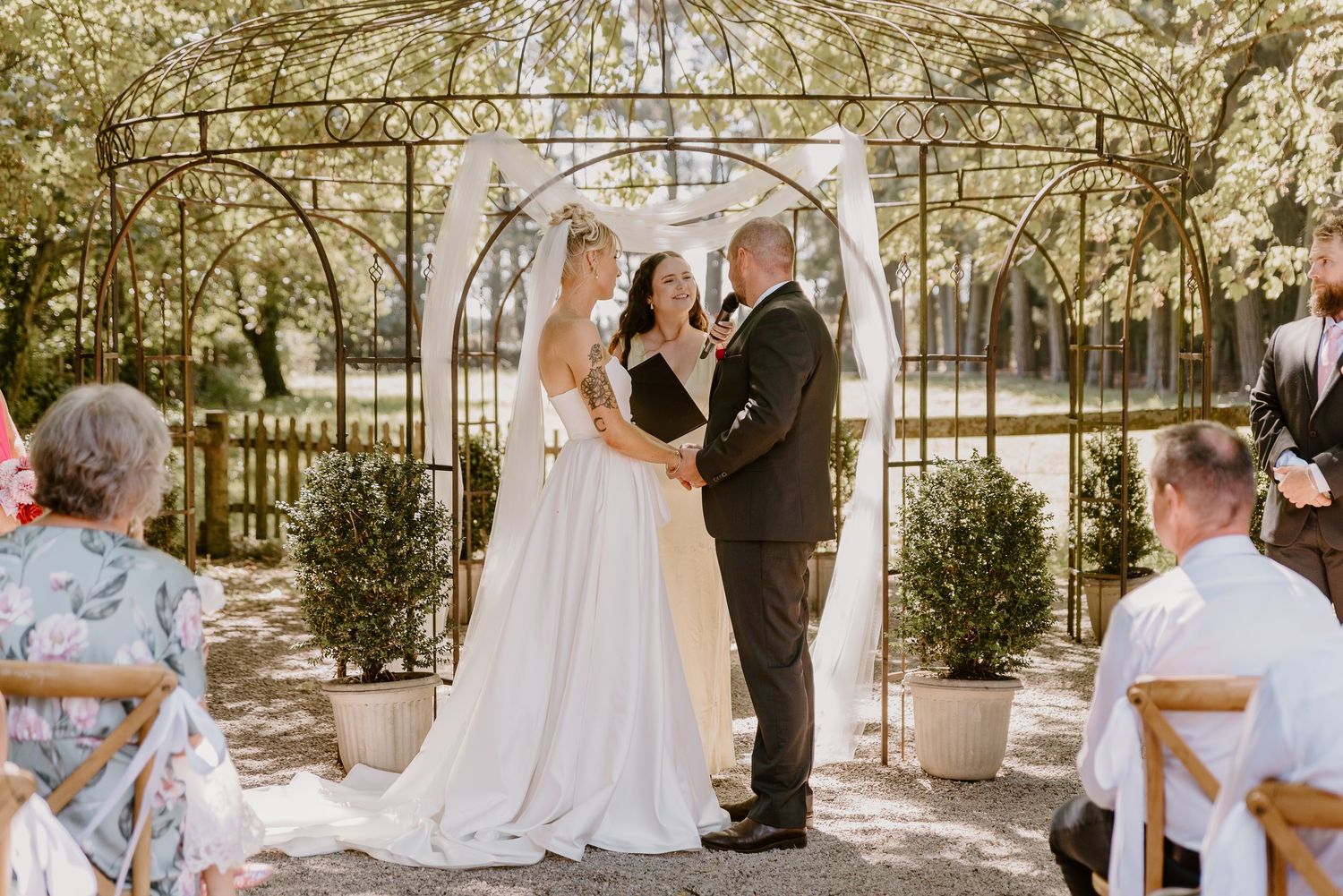 A wedding ceremony takes place under a white arch in a garden setting surrounded by greenery and twinkle lights.