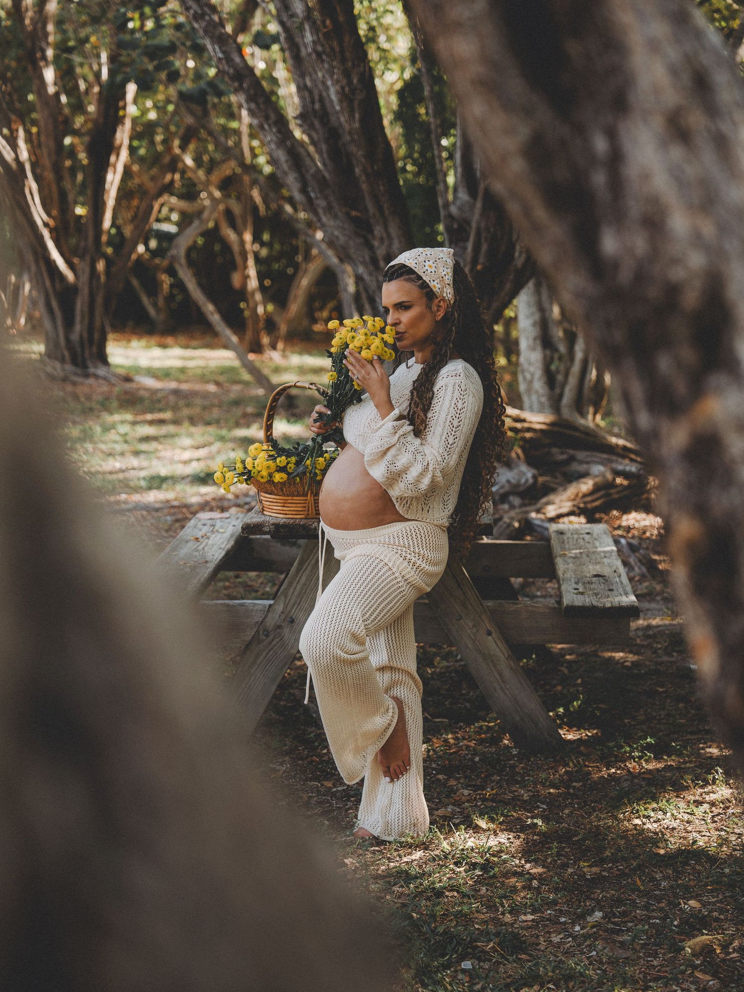 An expectant mother in white clothing poses with yellow flowers in a wooded setting.