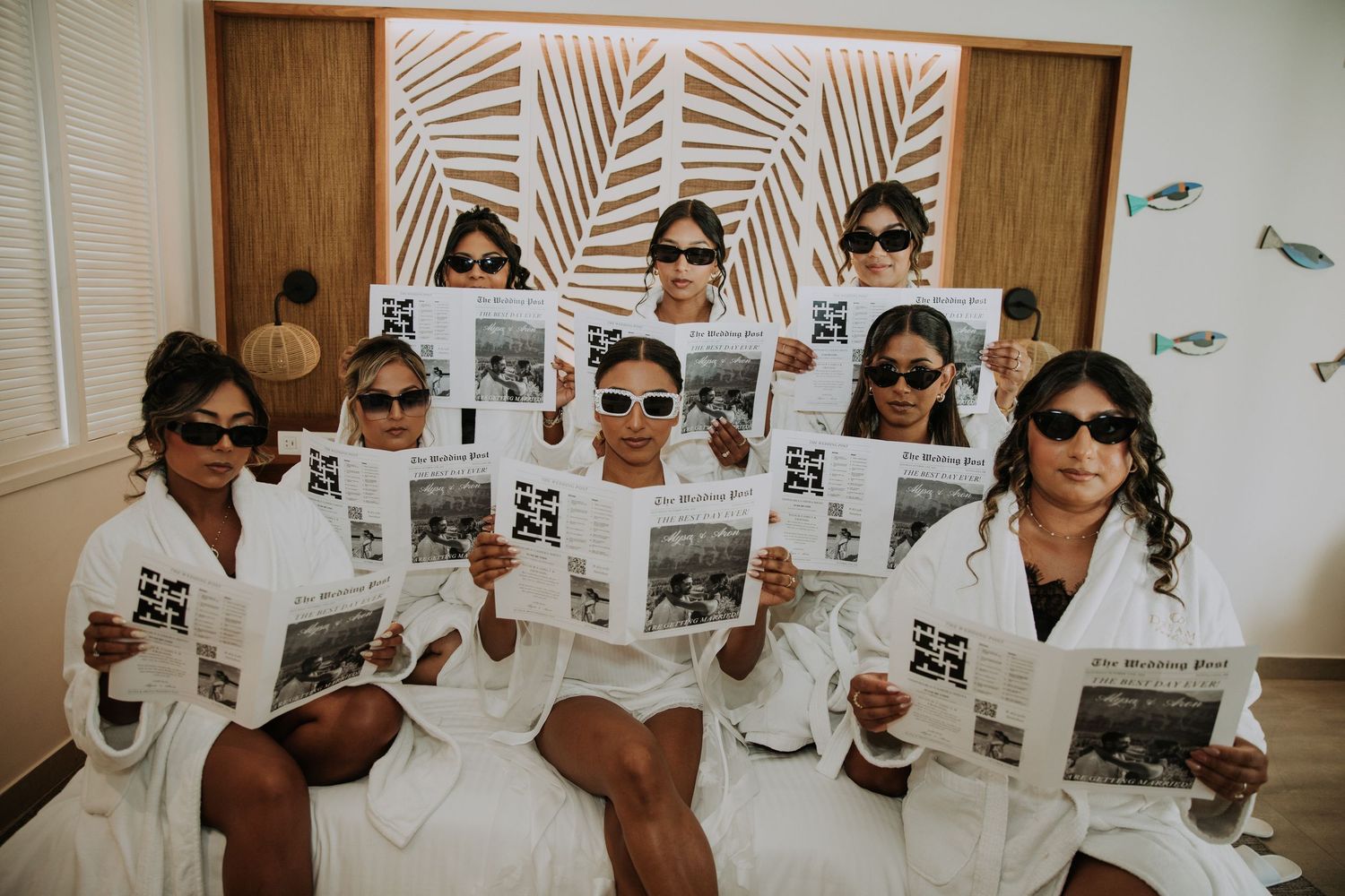 Bridesmaids wearing white robes and sunglasses pose with newspapers during wedding morning preparations.