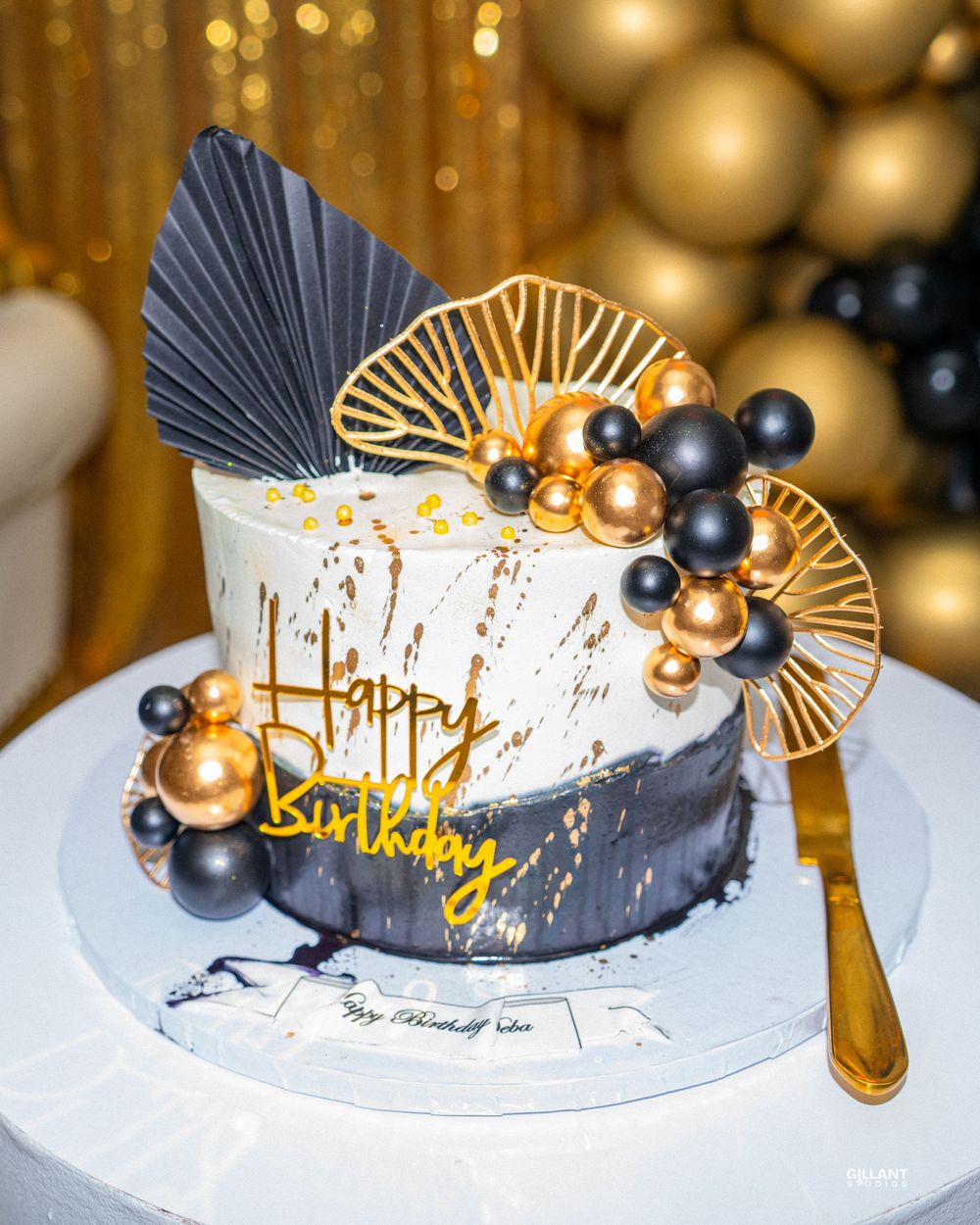 Modern birthday cake with black and gold decorations displayed against a warm bokeh background.