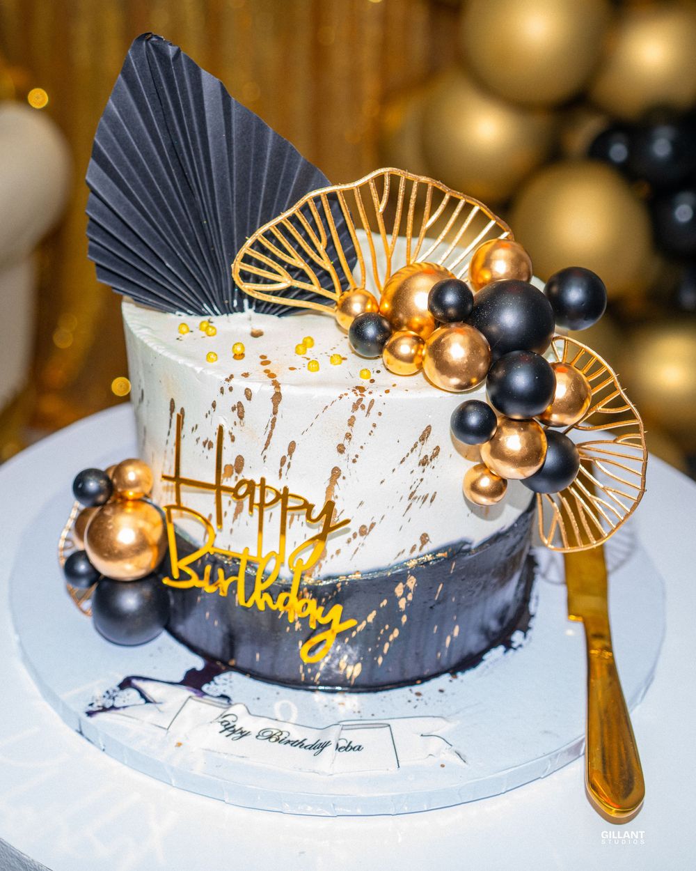 Elegant black and gold themed birthday cake with metallic balloon decorations and fan details.