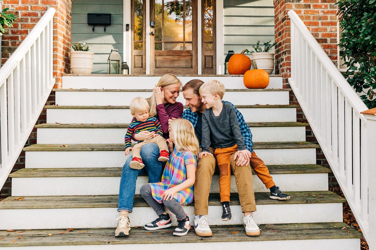 Family sitting together on Charlotte, NC home white porch steps with autumn pumpkin decoration in background.
