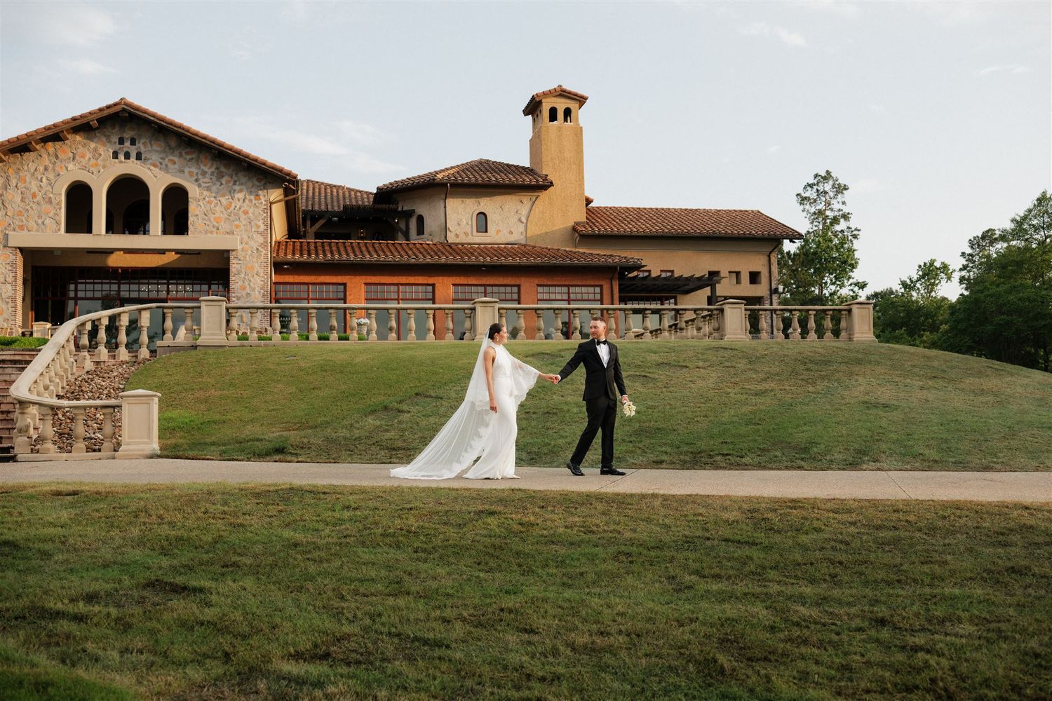 Wedding couple walking along elegant white stone archway surrounded by manicured garden and statues.