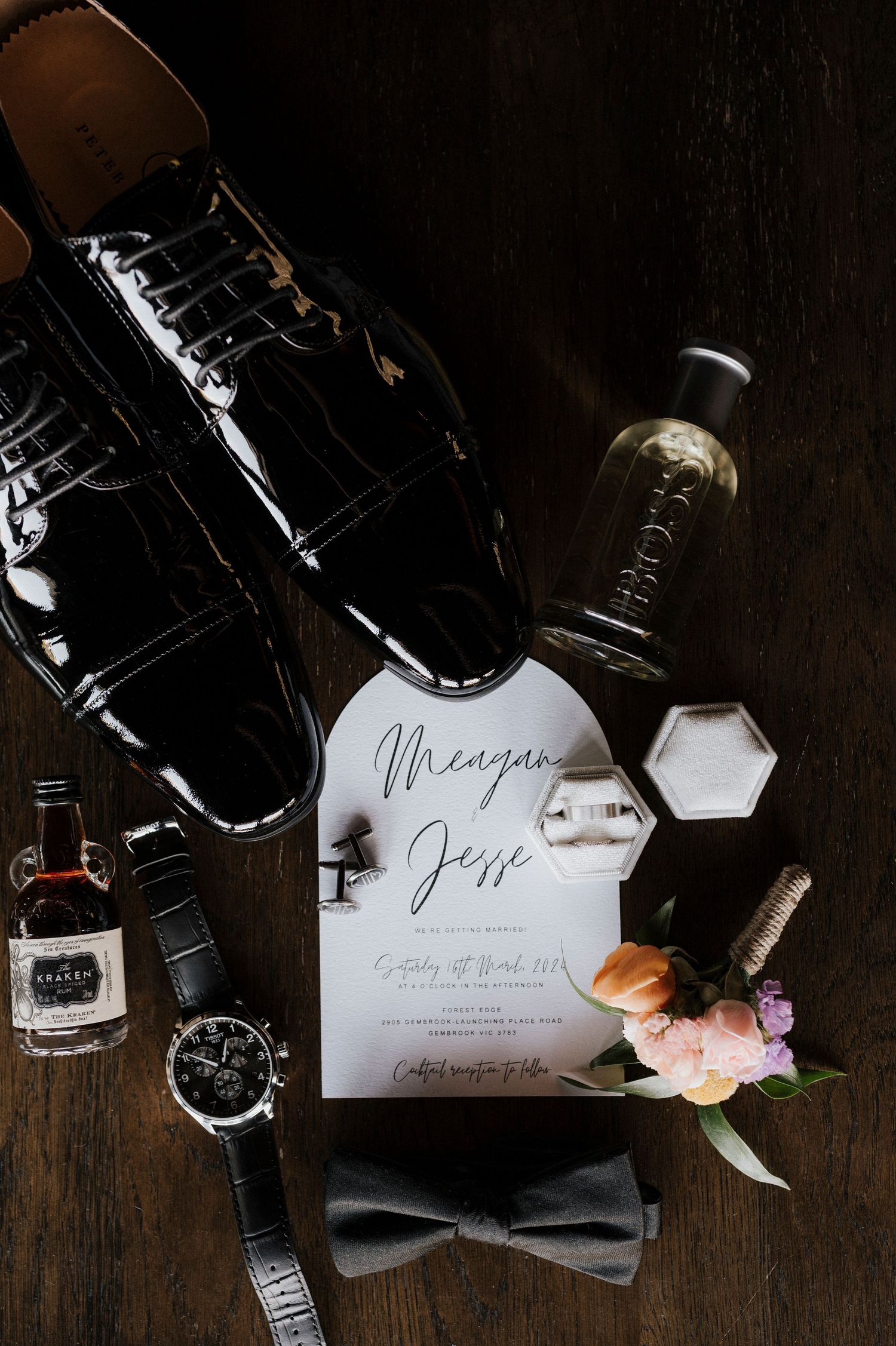 Wedding details flatlay on dark wood with invitations, watch, bow tie, and delicate pink flowers in elegant composition.
