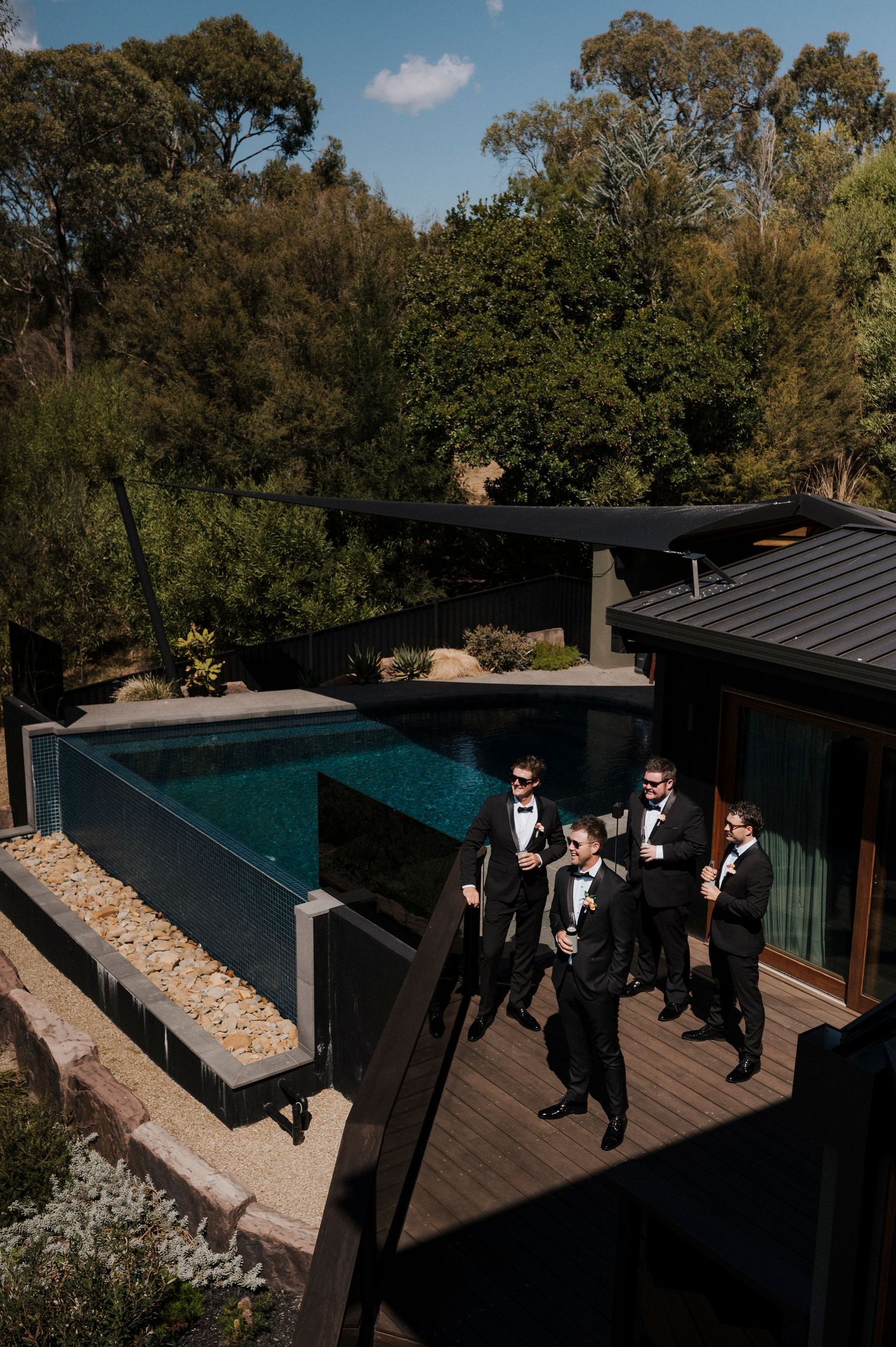 A group in formal attire stands on a modern deck with an infinity pool and lush trees in the background.