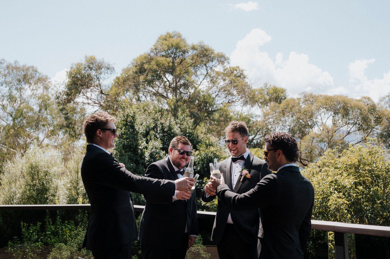 A group in formal black suits toast champagne together on an outdoor balcony surrounded by green trees.