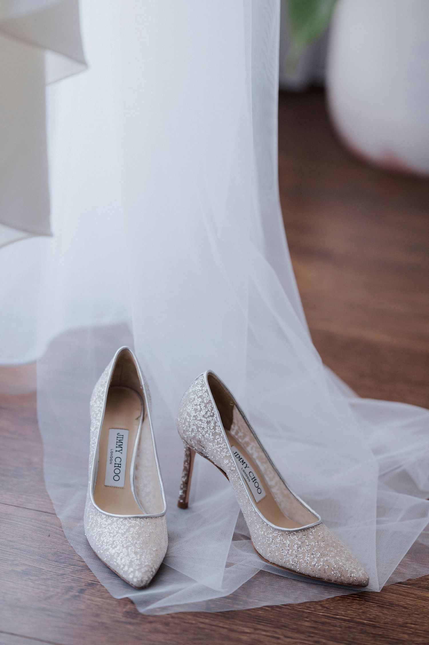 Elegant bridal shoes with intricate lace detailing rest beside flowing white tulle on a wooden floor.