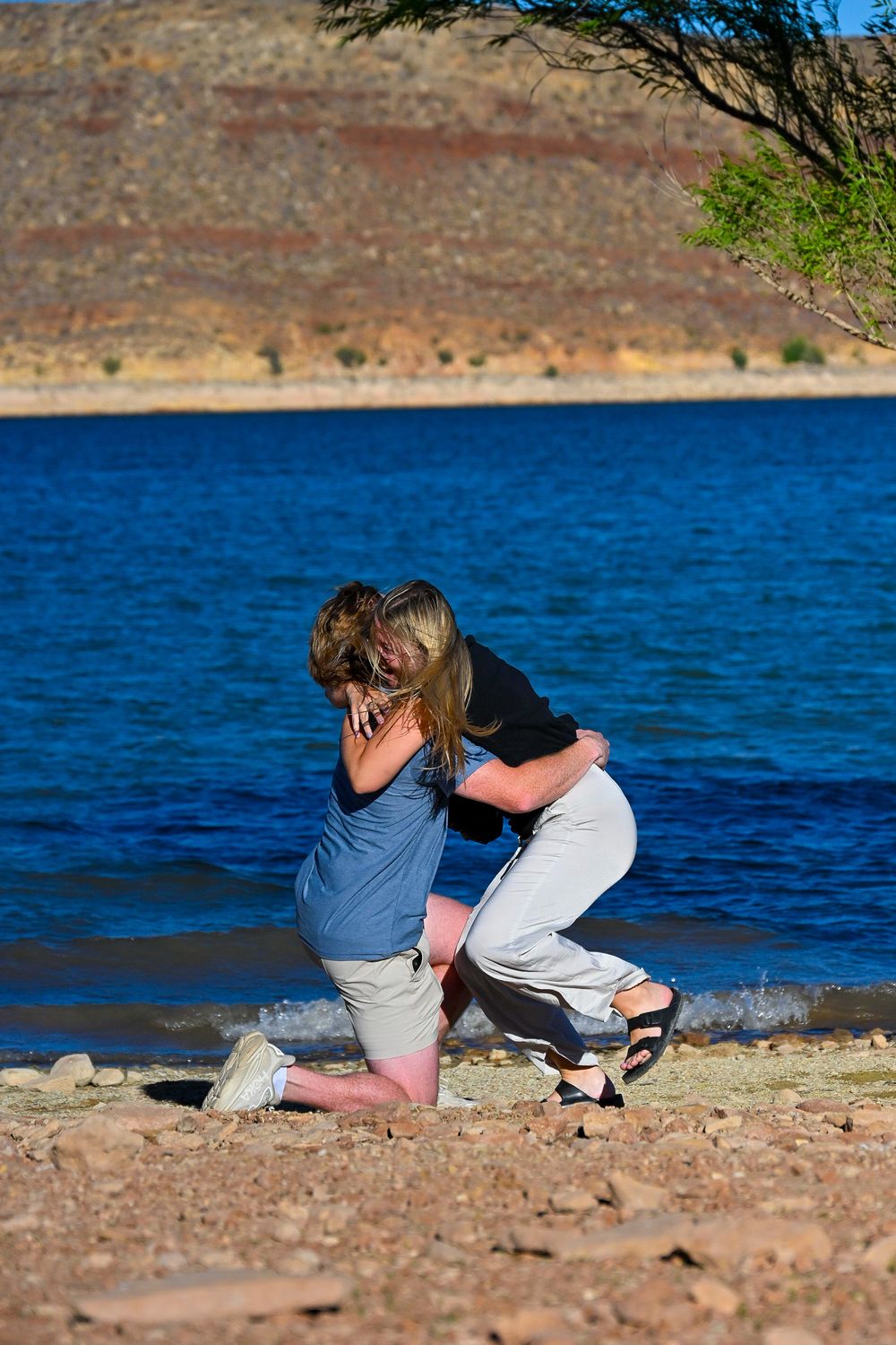 Couple shares emotional hug kneeling together on rocky lakeshore after a heartfelt marriage proposal outdoors.
