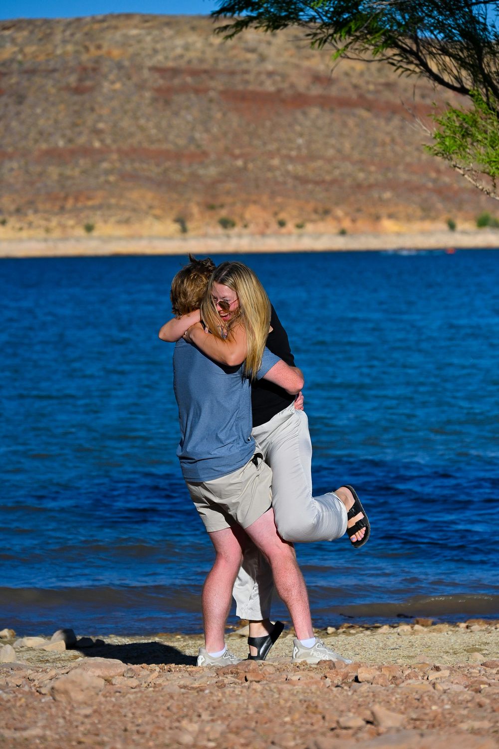 Joyful couple embraces and spins together on rocky beach beside vivid blue lake after a marriage proposal.
