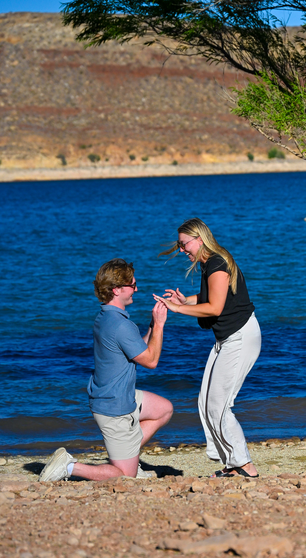 Man proposing on one knee to a woman on a sunny rocky beach beside a blue lake.