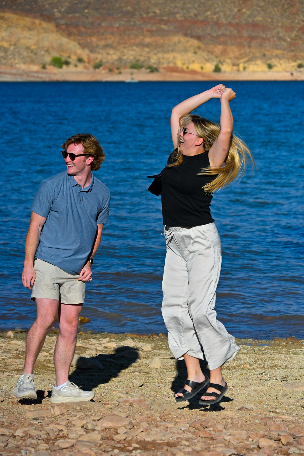 A man in sunglasses watches a woman dancing joyfully by a blue lake with rocky shores.
