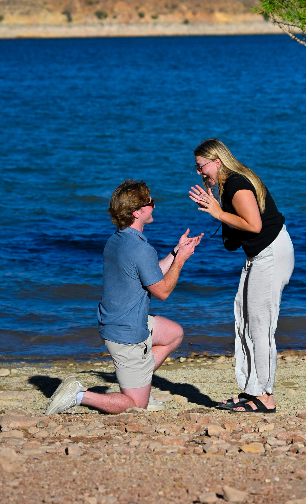A man proposes on bended knee as a woman reacts with excitement on a lakeside beach.