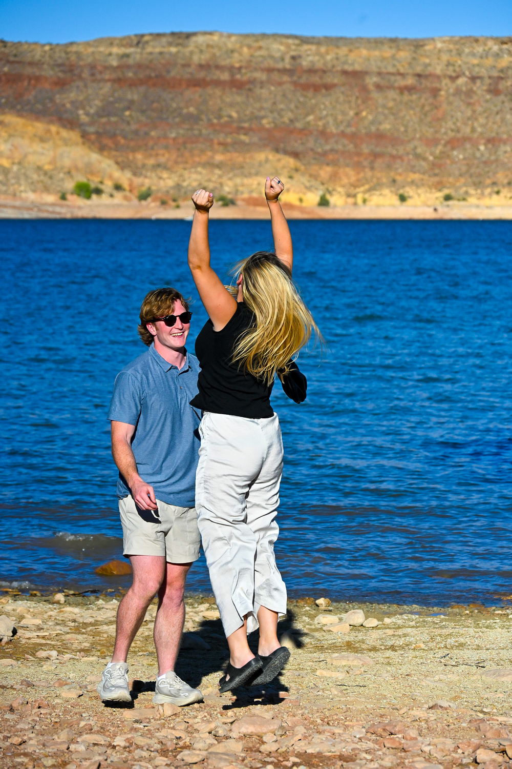 A woman celebrates with arms raised while a smiling man watches near a scenic blue lake.