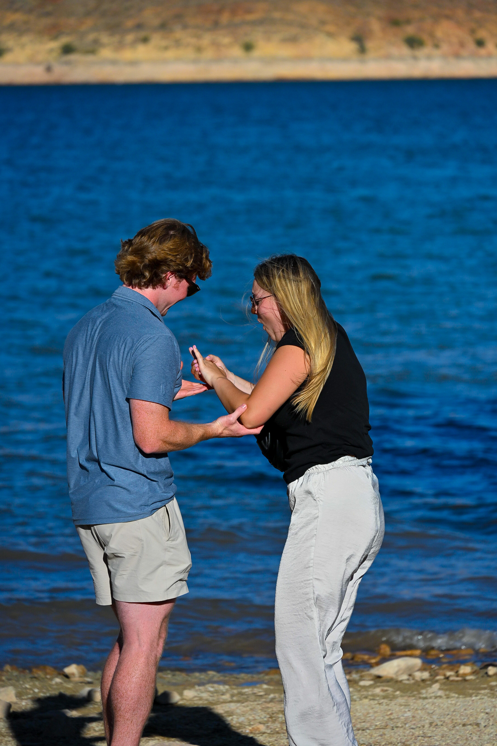 Couple sharing a joyful moment examining an engagement ring on a sunny lakeside shore.