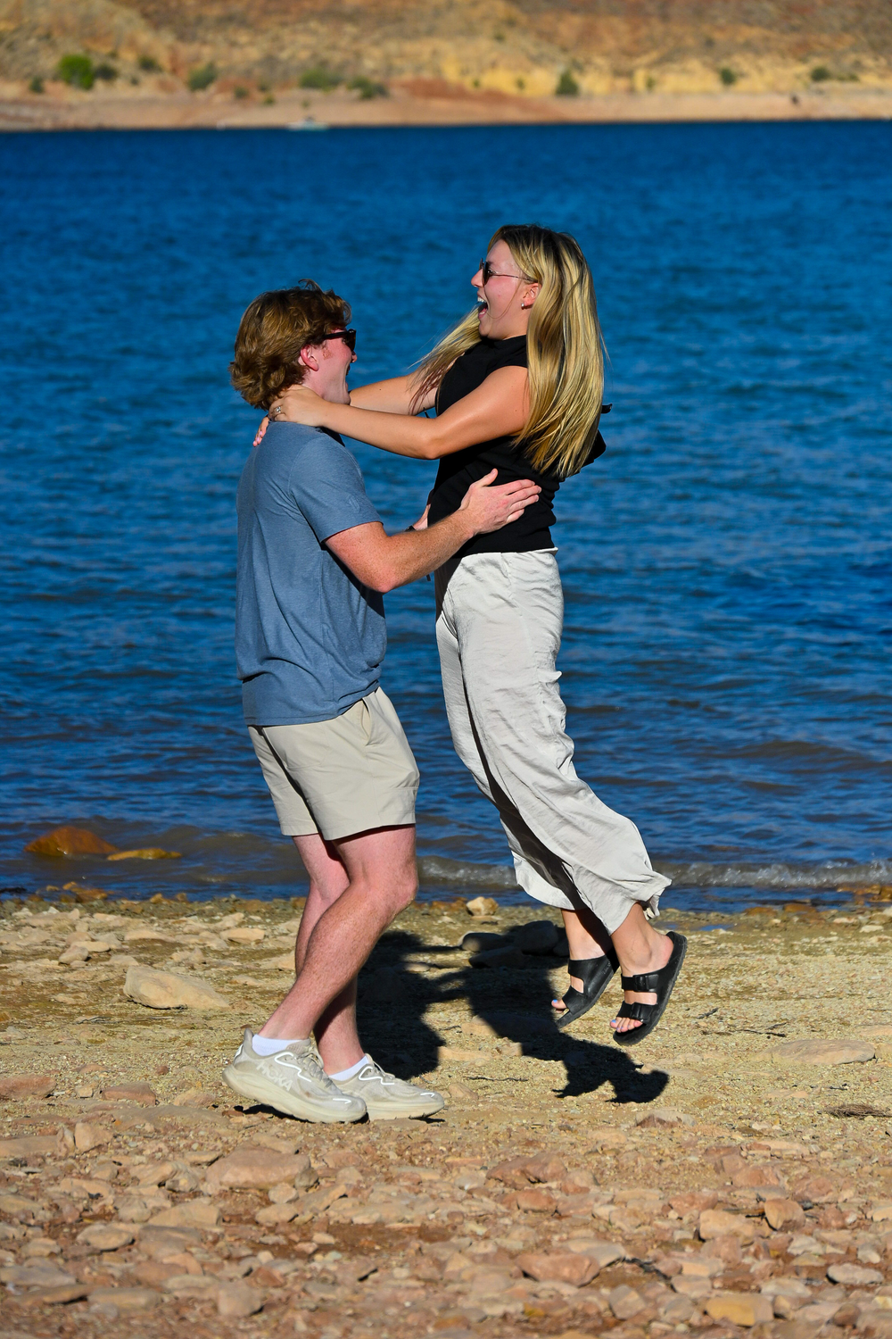 Joyful couple embracing and dancing together on a rocky shoreline beside blue water.