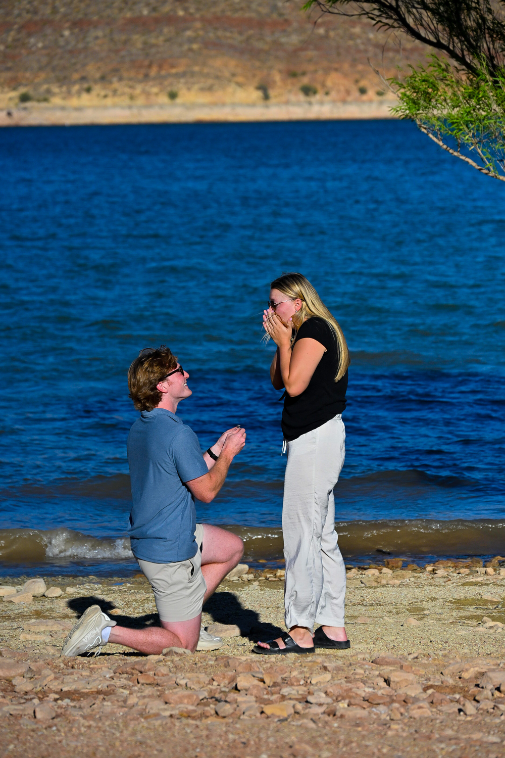 Man proposing on one knee to an emotional woman on a scenic rocky lakeside beach.