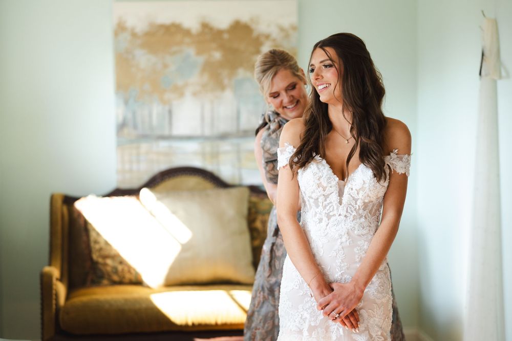 Mom and daughter in wedding attire share a tender moment in a sunlit room with soft lighting and elegant decor.