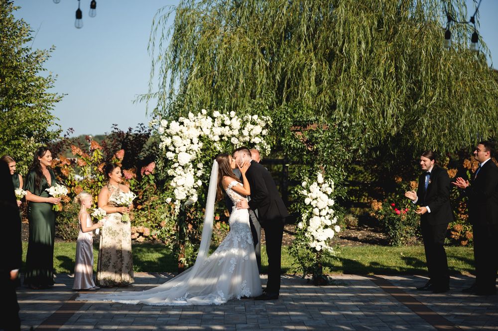 Couple's first kiss surrounded by a pergola with white roses during a golden hour ceremony at The Barn at Villa Venezia Middletown.