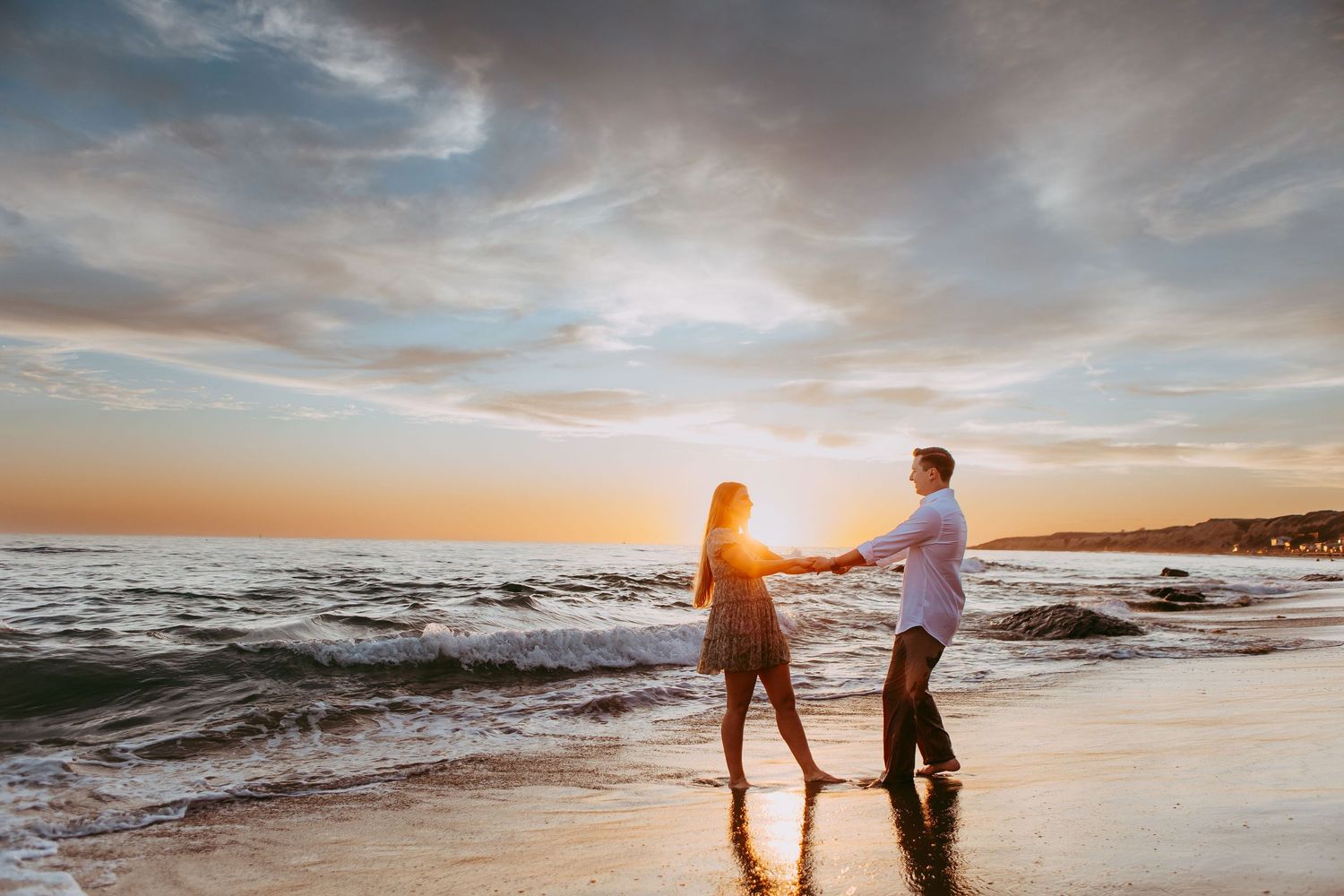 Silhouetted couple stand on sandy beach at sunset as waves crash along the shoreline during engagement session in Orange County, California.