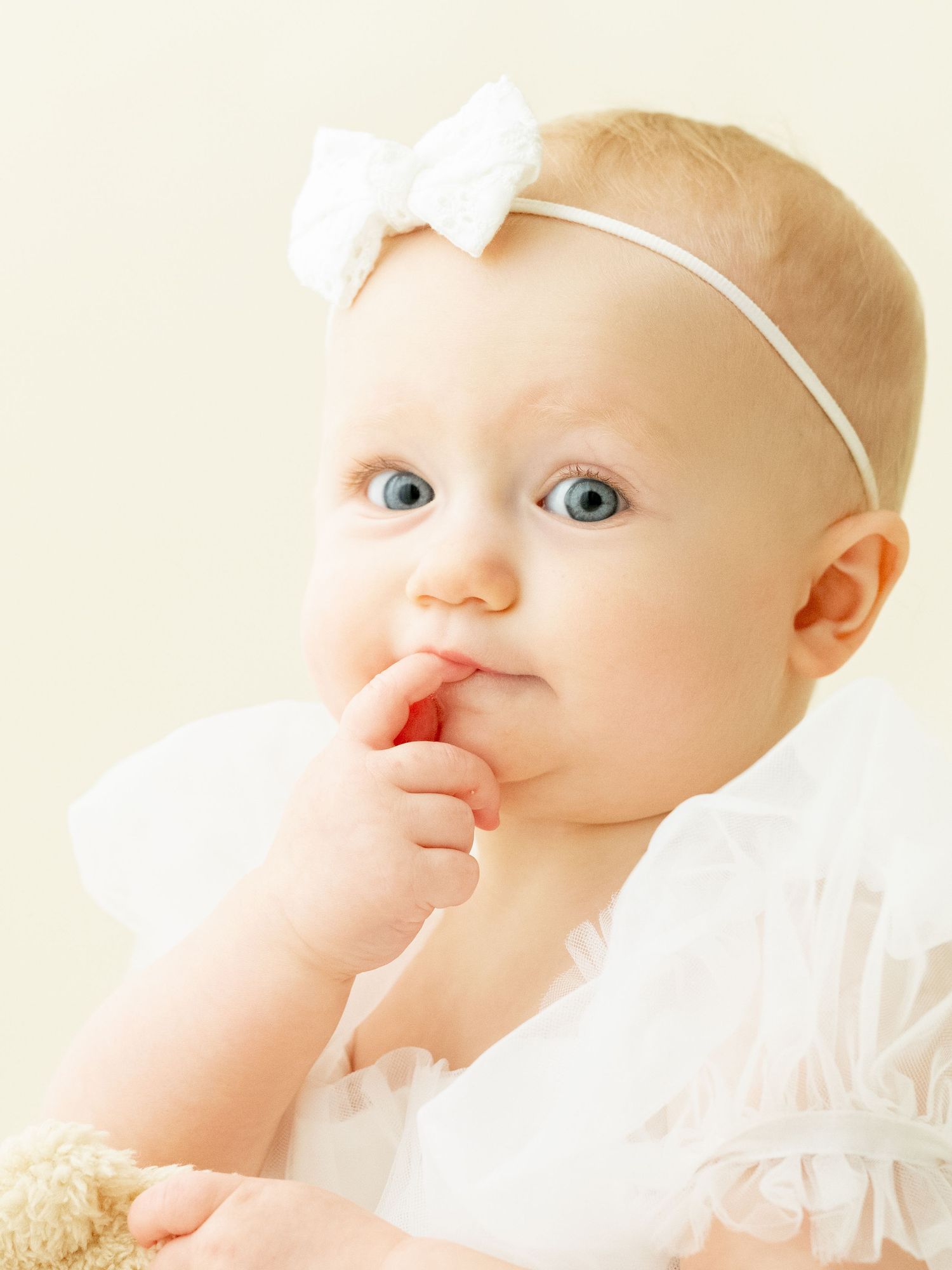 Baby in white outfit wearing delicate white headband poses for series of sweet expressions against light backdrop.