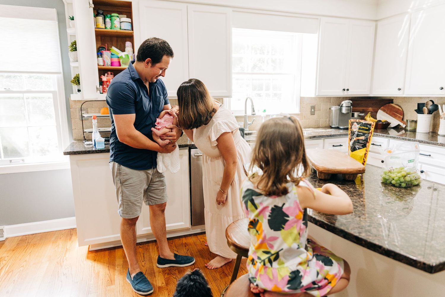 A family gathered together in a bright kitchen with hardwood floors and white cabinets during a casual moment at home.