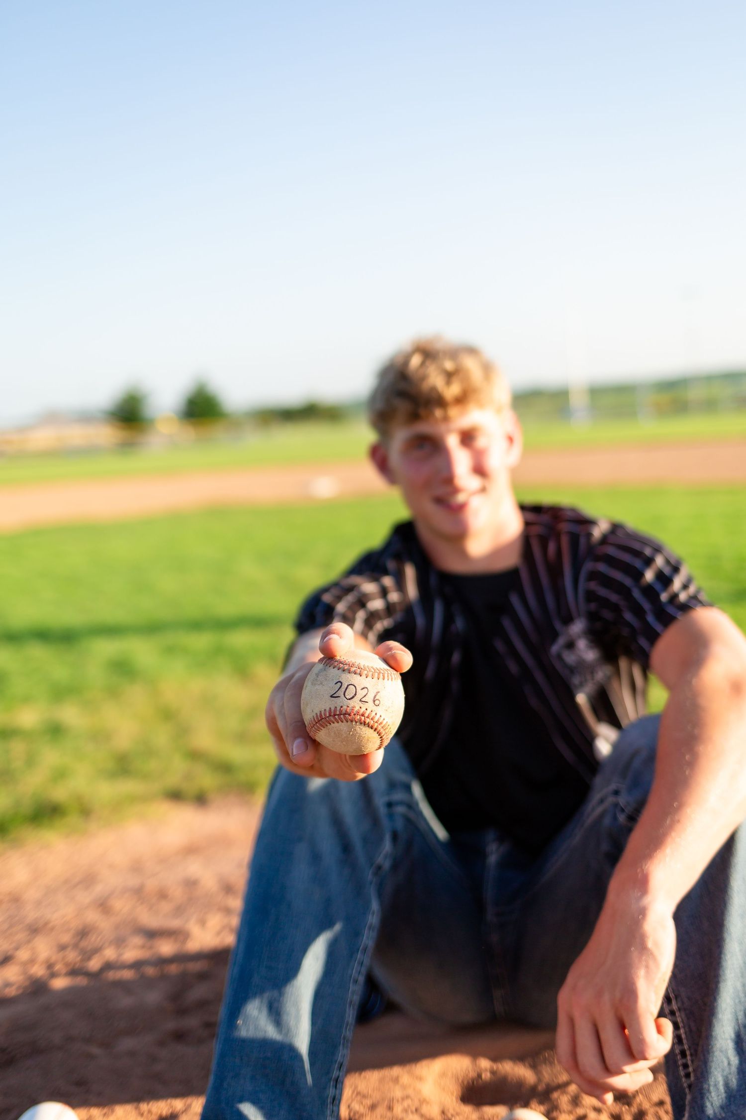 A person sits in a baseball field at sunset holding a baseball while wearing jeans and a dark shirt.