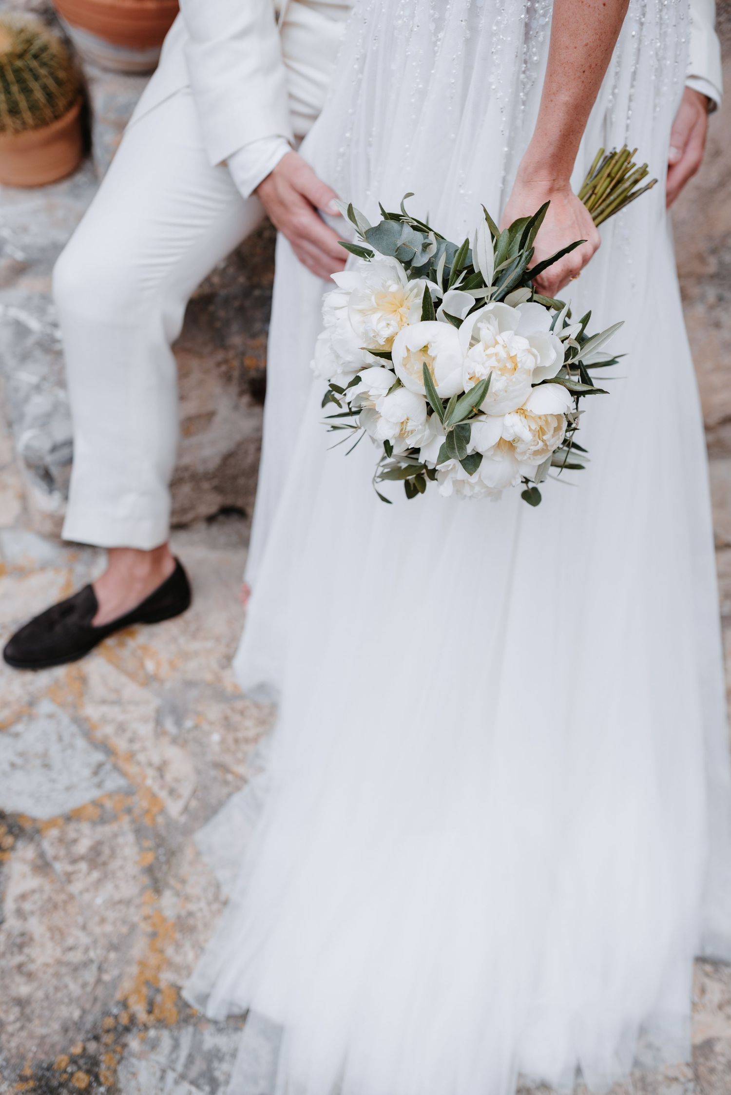 A delicate white floral bridal bouquet with eucalyptus held against a flowing tulle wedding dress and stone floor.