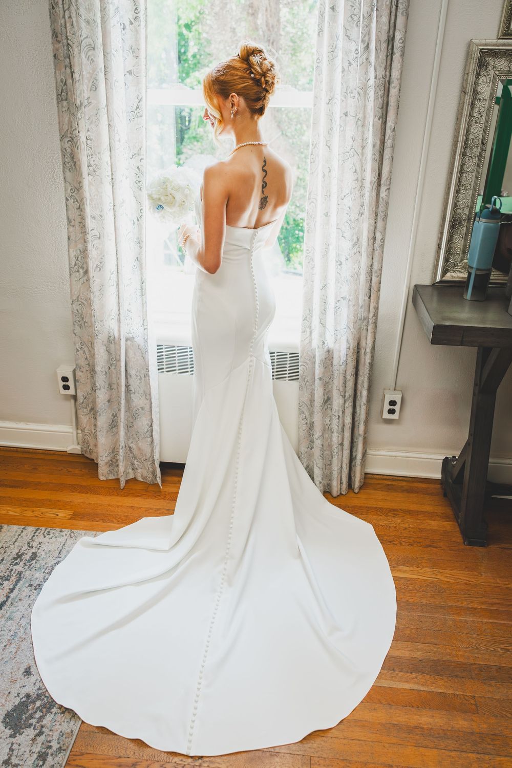 Elegant white wedding gown with dramatic train photographed from behind on hardwood floor by lace curtained window.