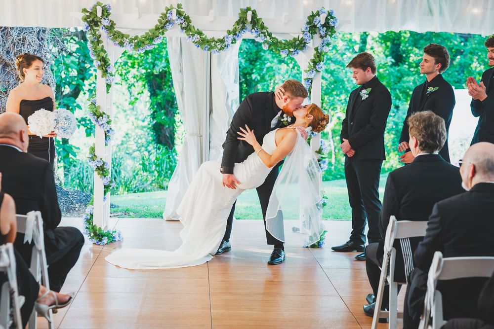 A wedding reception panorama shows guests seated at tables while a couple dances in a room decorated with green foliage.