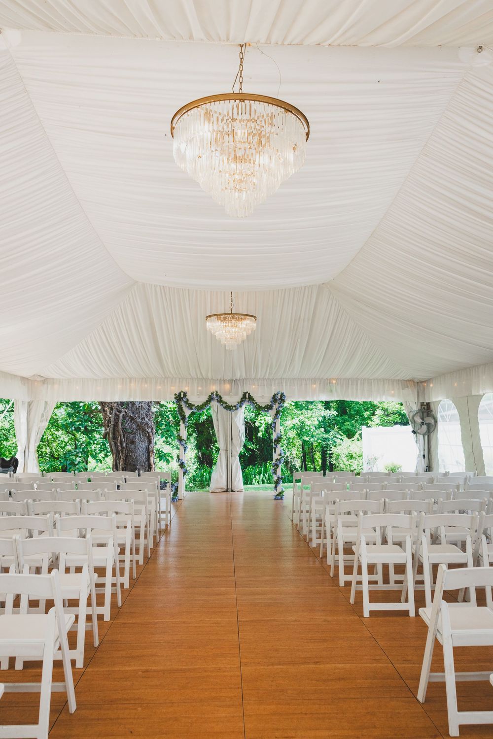 Elegant wedding ceremony tent with crystal chandeliers, white draping, and rows of white chairs on a wooden floor.