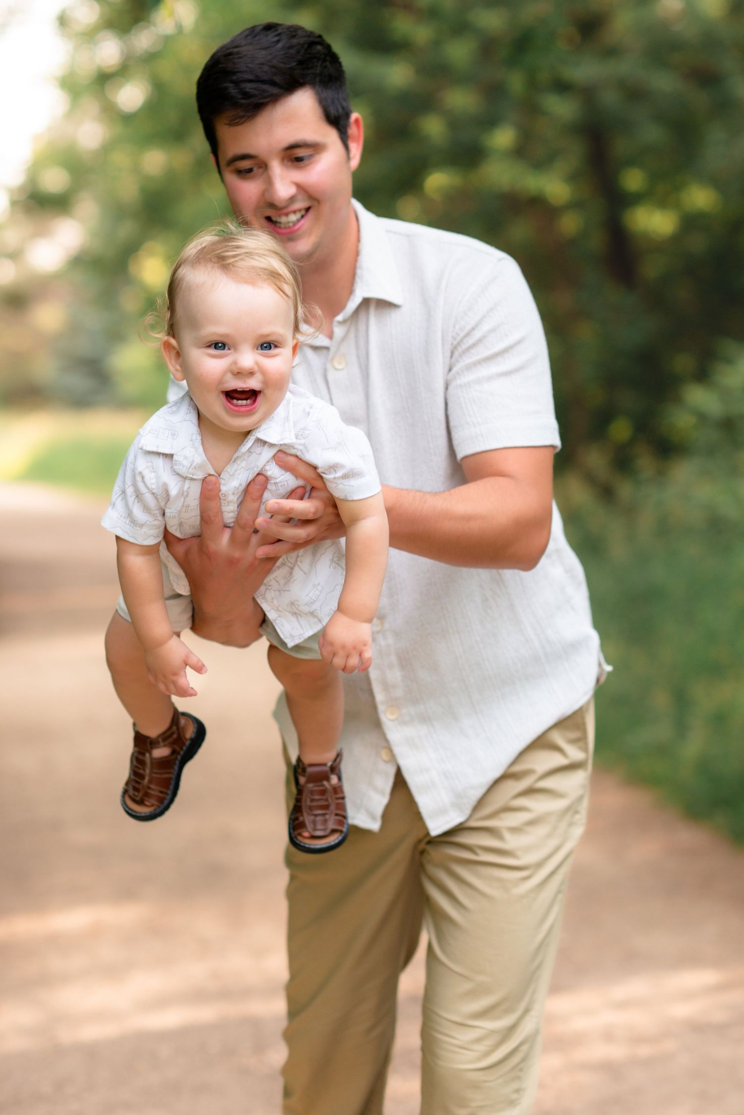 A joyful moment captured on a wooded path as parent lifts laughing toddler in matching white shirts outdoors.