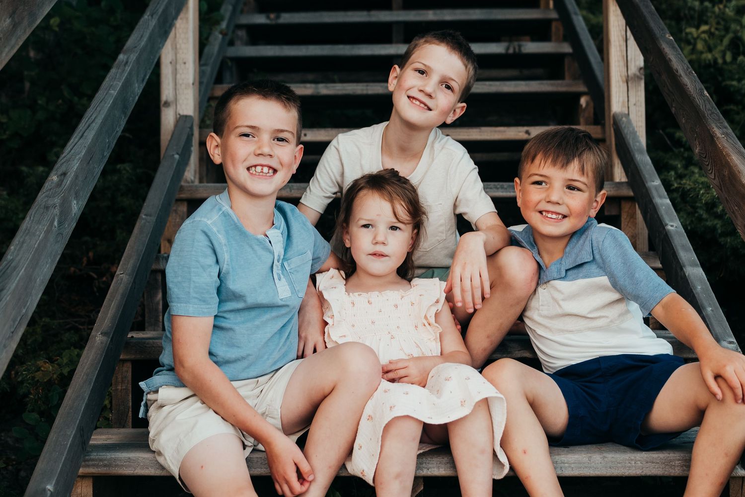 Children wearing casual clothing sitting together on wooden stairs outdoors smiling at the camera.