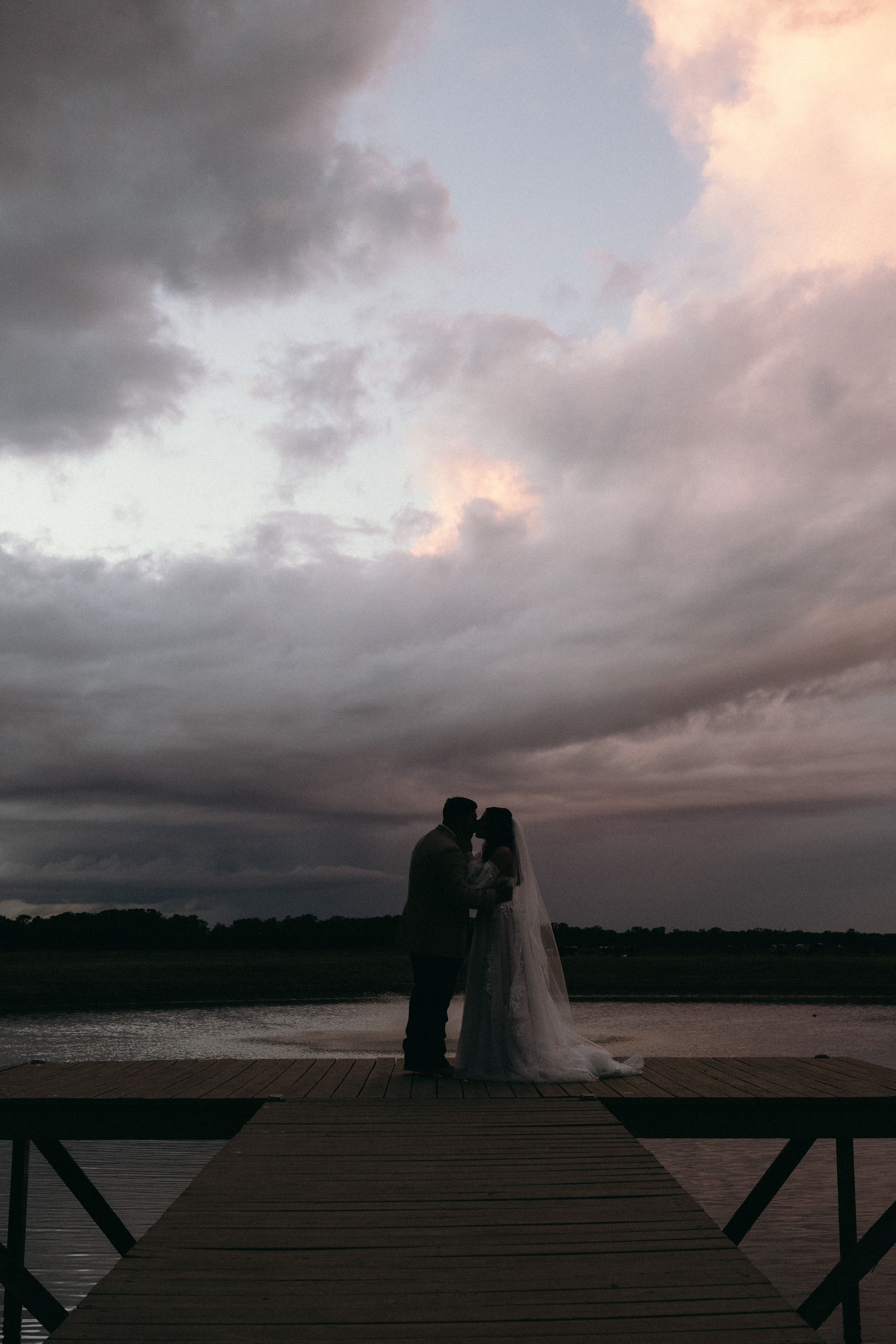 Silhouetted couple shares romantic moment on wooden dock at sunset with dramatic storm clouds overhead.