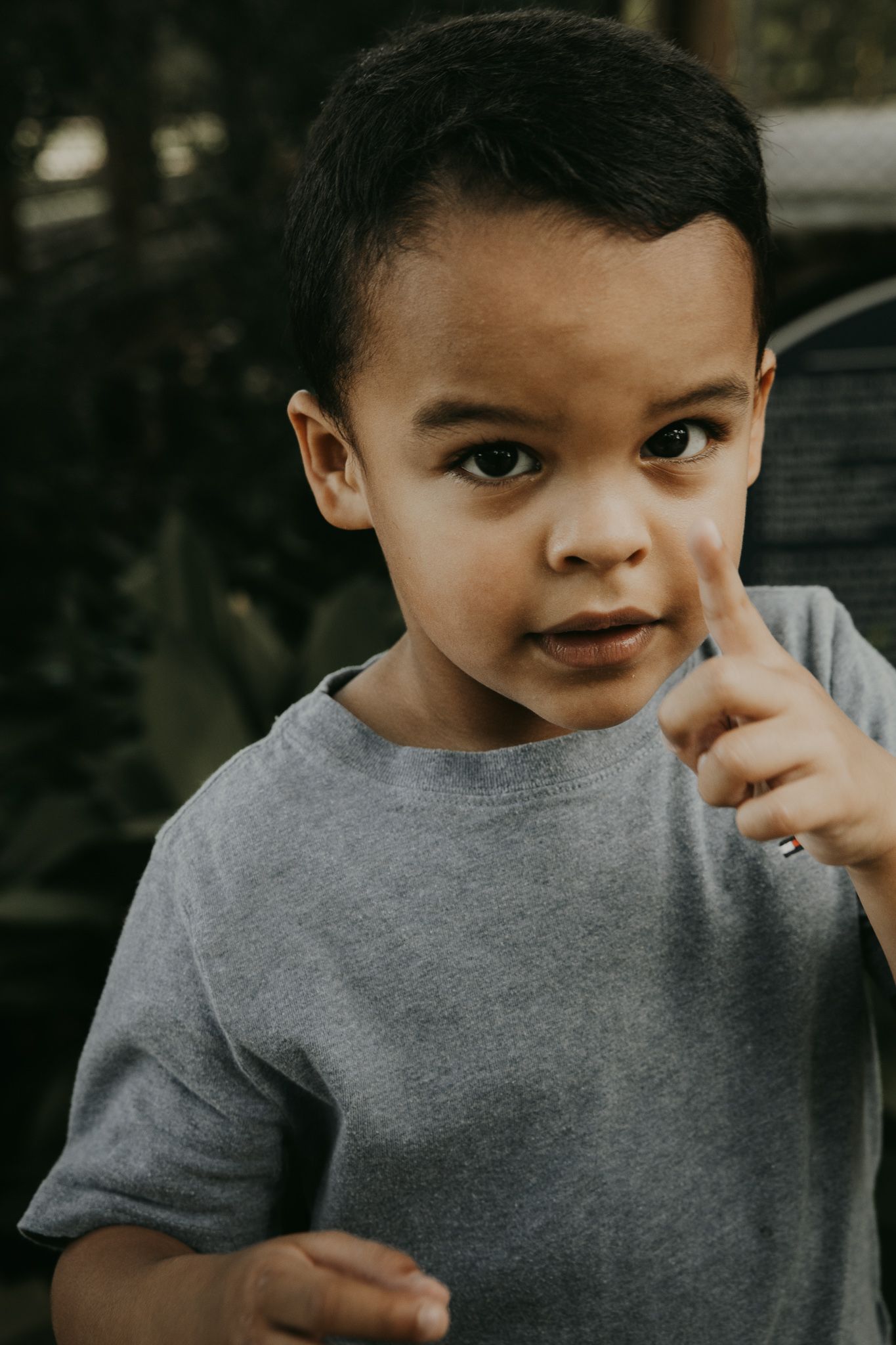 Young child in a gray t-shirt holds finger to their face in a thoughtful expression against blurred outdoor background.
