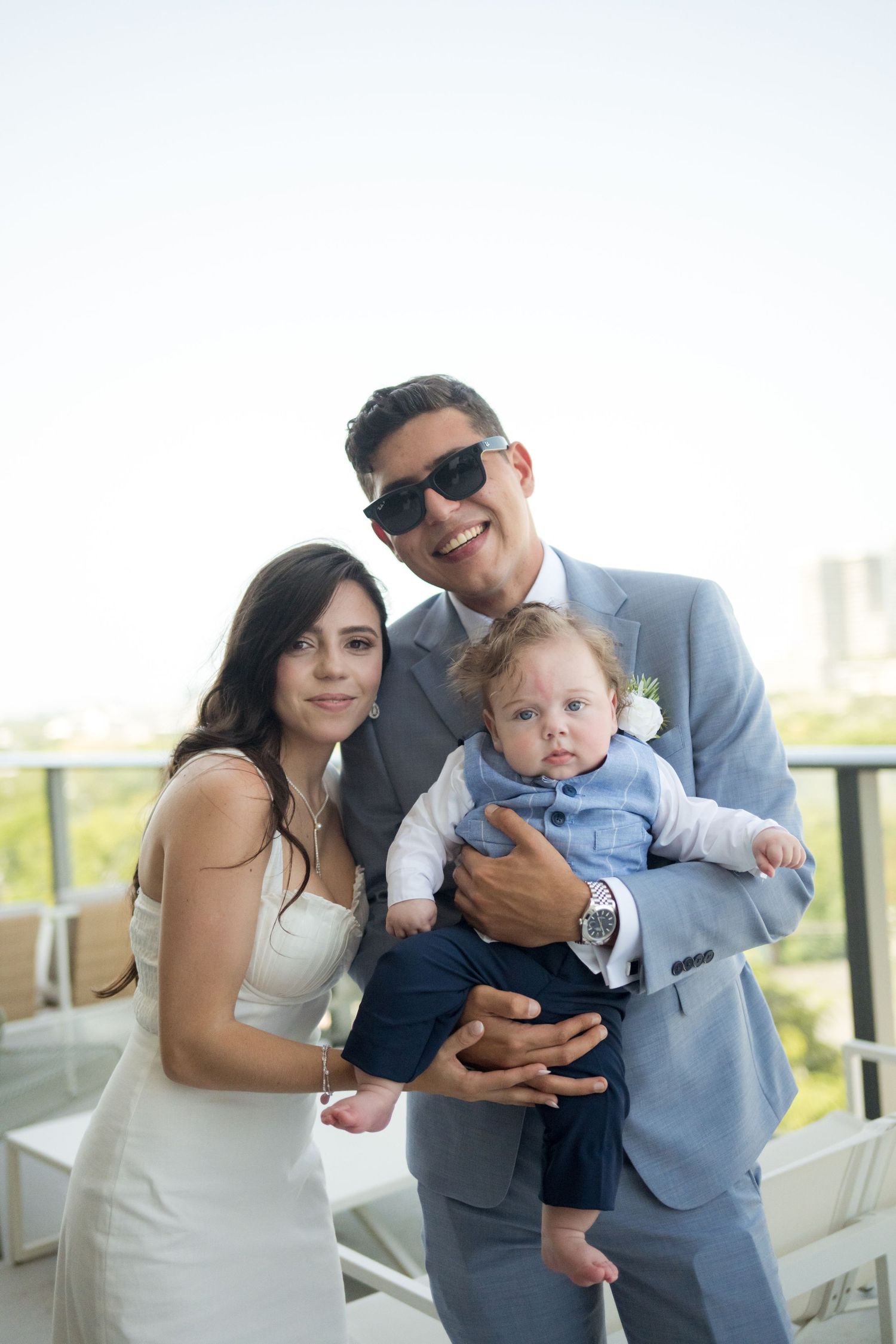 Happy couple in formal attire poses outdoors with a baby in blue outfit on a sunny day.
