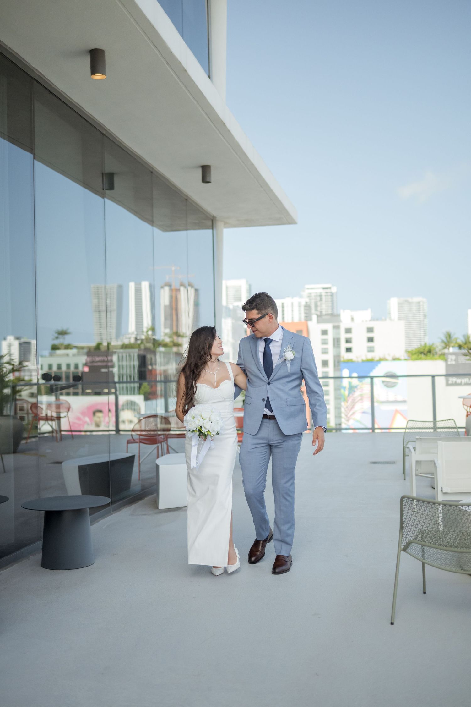 Couple in wedding attire walking on modern rooftop terrace with city skyline view in background.
