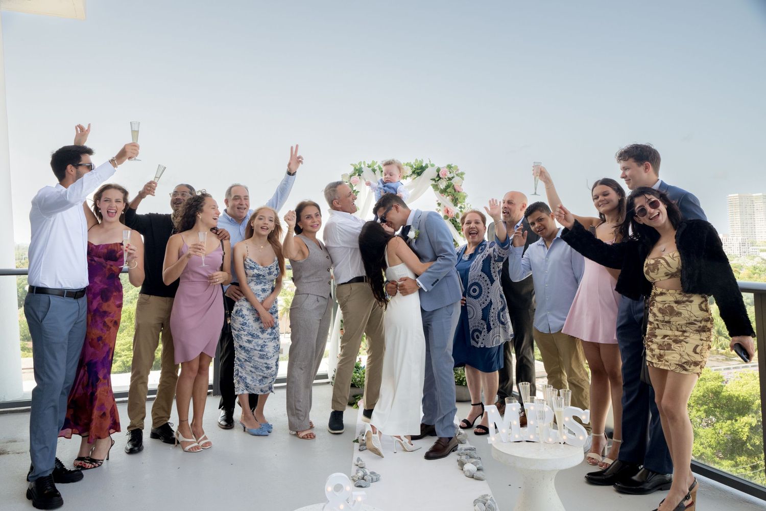 A group celebrates joyfully during a daytime outdoor wedding celebration with drinks and toasts against a bright sky.