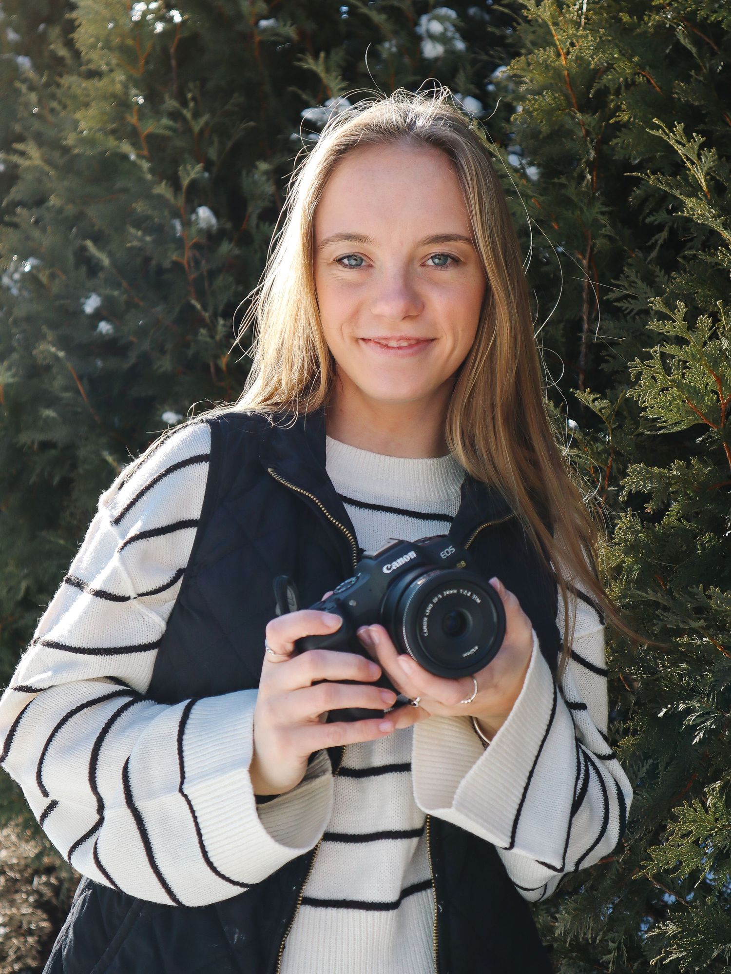 A photographer smiles while holding a camera outdoors against an evergreen backdrop.
