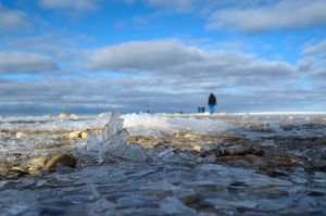 Wave splashes over ice formations on a beach with a distant figure silhouetted against cloudy sky.