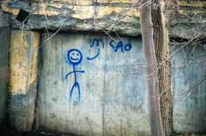 Simple blue graffiti stick figure with a smiley face drawn on a weathered concrete wall.