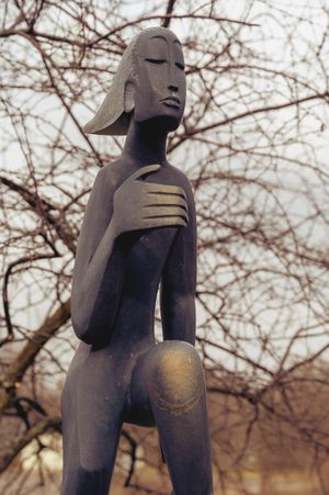 Modern abstract metal sculpture of a stylized figure in contemplative pose against bare branches.