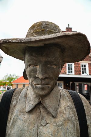 Close-up view of a weathered stone statue wearing a wide-brimmed hat in front of a brick building.