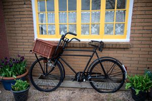 Vintage black bicycle with brown wicker basket parked against brick wall beneath yellow window and purple flowers.