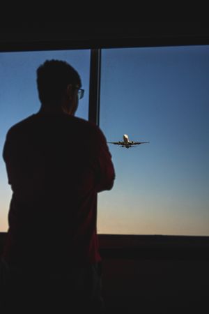 Silhouetted passenger watching commercial airplane approach through airplane window during sunset.