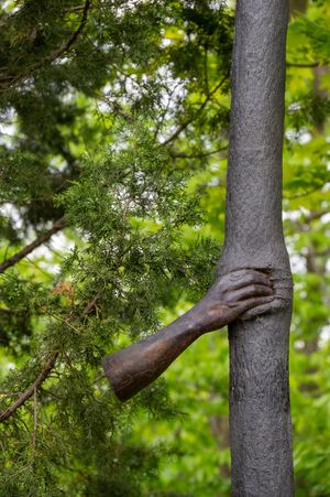 Bronze sculpture of an arm grasping a tree trunk against lush green foliage in the background.