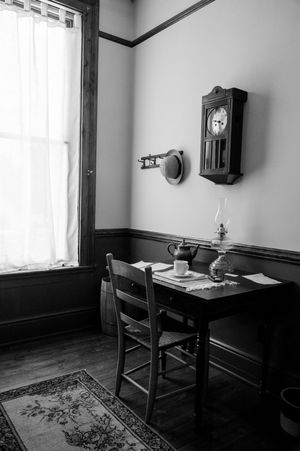 A vintage wooden desk with chair sits beneath a wall-mounted clock in a sparse room with hardwood floors and an area rug.